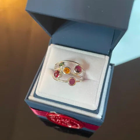 A silver-toned ring with five colorful gemstones (red, orange, green, and two pinkish-red) displayed inside an open blue jewelry box with a white insert.