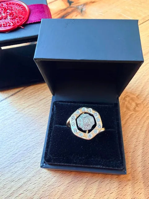 A gold-toned ring with a silver-toned shield emblem in the center and small clear stones around it, displayed in an open black jewelry box on a wooden surface.