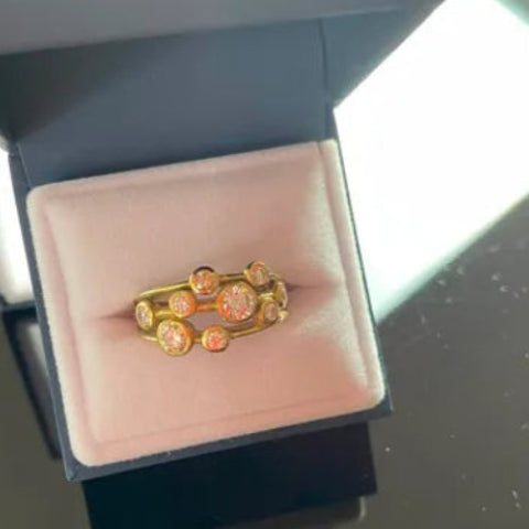 A gold ring with small clear stones and orange-gold accents, displayed in an open dark gray jewelry box with a pale pink cushion.