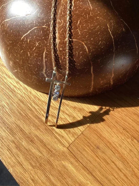 A silver cross pendant necklace with a small clear gemstone, hanging from a brown cord in front of a polished brown coconut shell on a wooden surface.
