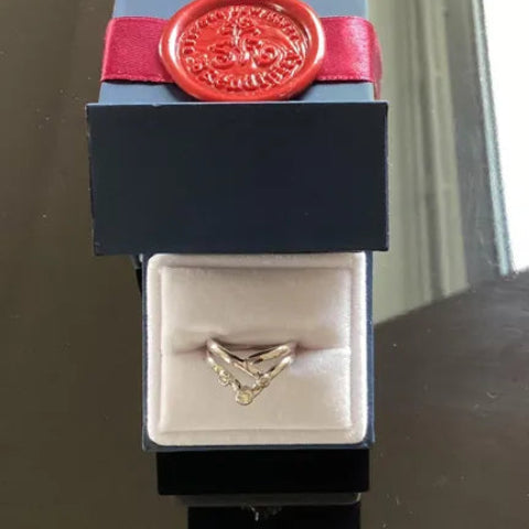 A gold ring with a V-shaped chevron design and small clear stones, displayed inside an open black jewelry box with a red wax seal on a dark reflective surface.