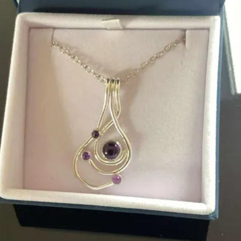 A gold pendant necklace with a teardrop-shaped design, featuring a central purple gemstone and two smaller purple accent stones, displayed inside an open jewelry box.