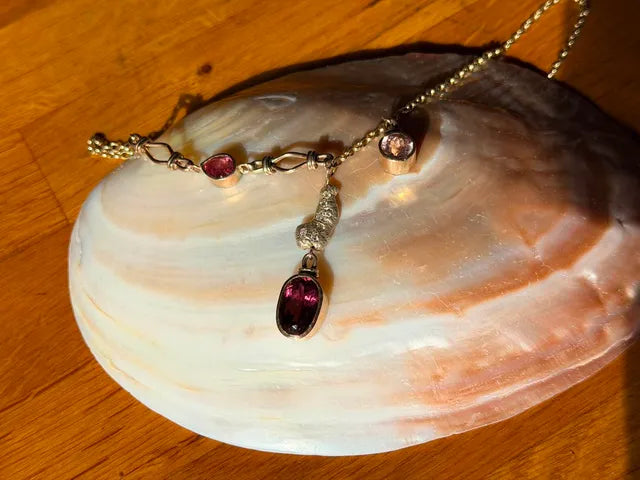 A gold-toned necklace with dark red gemstone accents and a dangling oval pendant, displayed on a large white clam shell over a wooden surface.