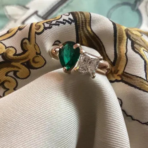 A close-up of a ring with a green marquise-cut gemstone and a silver-toned setting, resting on draped silk fabric with a gold and white baroque pattern.