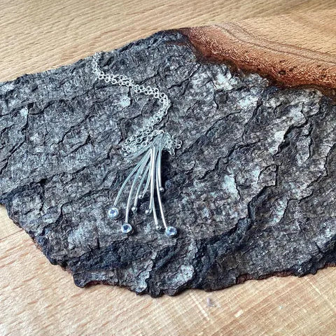 A silver-toned necklace with a delicate chain and multiple thin dangling strands, displayed on a textured piece of bark over a light wood surface.