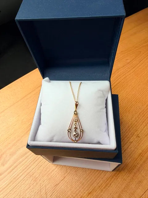 A gold teardrop pendant necklace displayed on a white cushion inside an open navy blue jewelry box, set on a light wood tabletop.