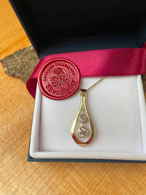 A gold teardrop pendant necklace with two clear round-cut stones, presented in an open white jewelry box with a dark blue base, a bright pink ribbon, and a red wax seal.