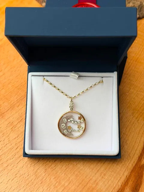 A gold-tone pendant necklace with a circular charm featuring a crescent moon and small star details, displayed inside an open dark blue jewelry box on a wooden surface.