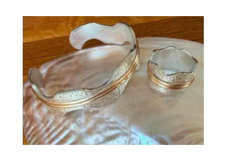 A mother-of-pearl shell dish with iridescent pearly tones and silver-toned metal trim, holding a small matching silver ring holder, photographed on a wooden surface.