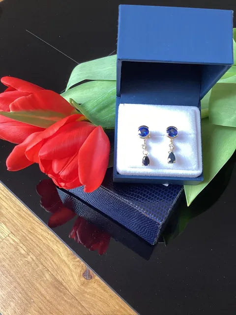 A pair of drop earrings displayed in an open blue jewelry box on a white insert, placed on a dark reflective surface with red tulips and green leaves around it.