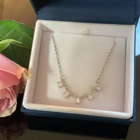 A silver necklace with small square-cut clear stones is displayed inside an open dark blue jewelry box with a white insert. A pink rose is partially visible on the left side of the box.