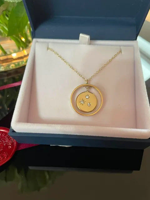 A gold pendant necklace with a double-ring design and small star details, displayed inside an open navy blue jewelry box with a light pink velvet insert.